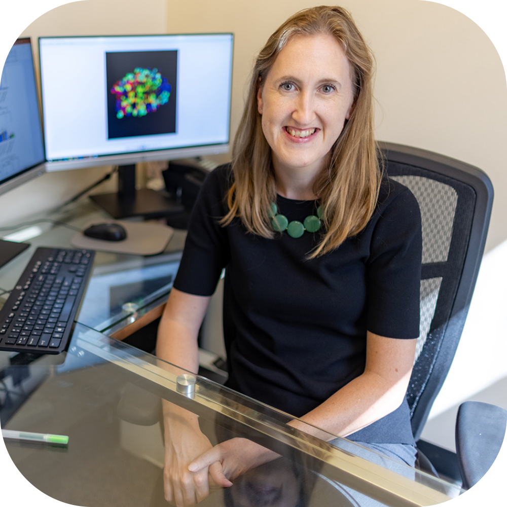 Smiling woman at glass desk in office, portrait photograph with computer monitors