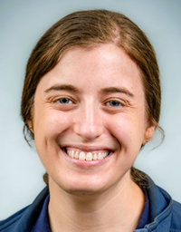 Smiling young woman headshot, brown hair pulled back, navy top, gray background