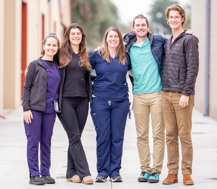 Five people stand together, smiling on a broad walkway with buildings in the background.