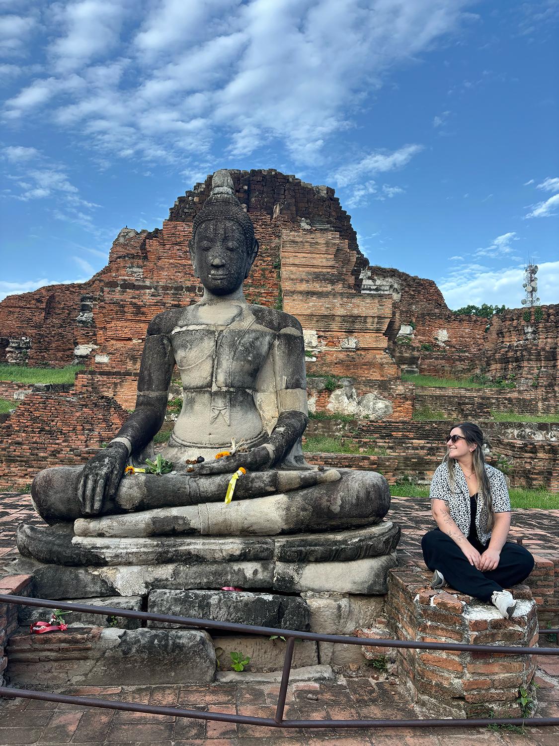 A person sitting next to a much larger sculpture of a god figure in a set of ruins.