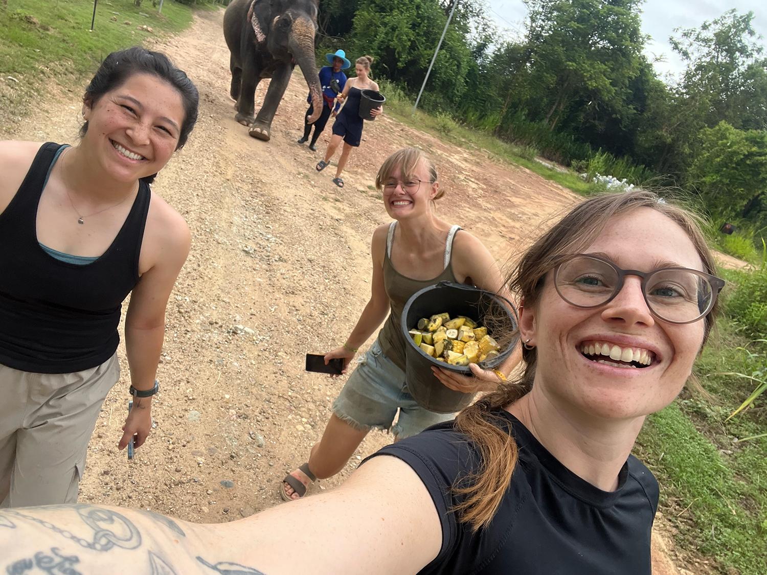 Three people taking a selfie, holding buckets of food with an elephant and people in the background.