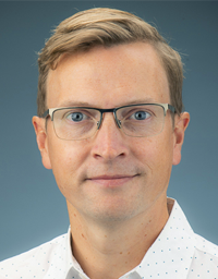 Headshot photograph of a man with glasses, light brown hair, white shirt, blue-gray background