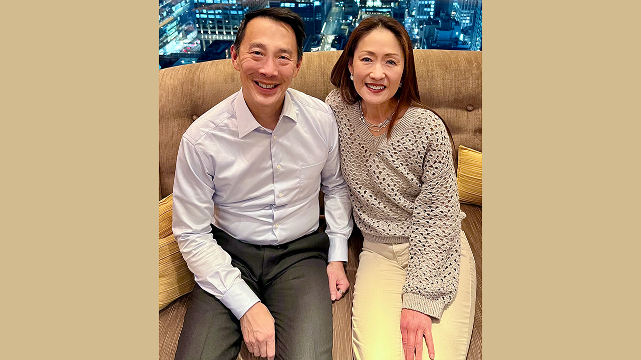 Ken and Kathy sitting on a couch with the night city skyline in the background window.