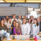 Group of smiling laboratory researchers in coats gathered around a cluttered lab bench.